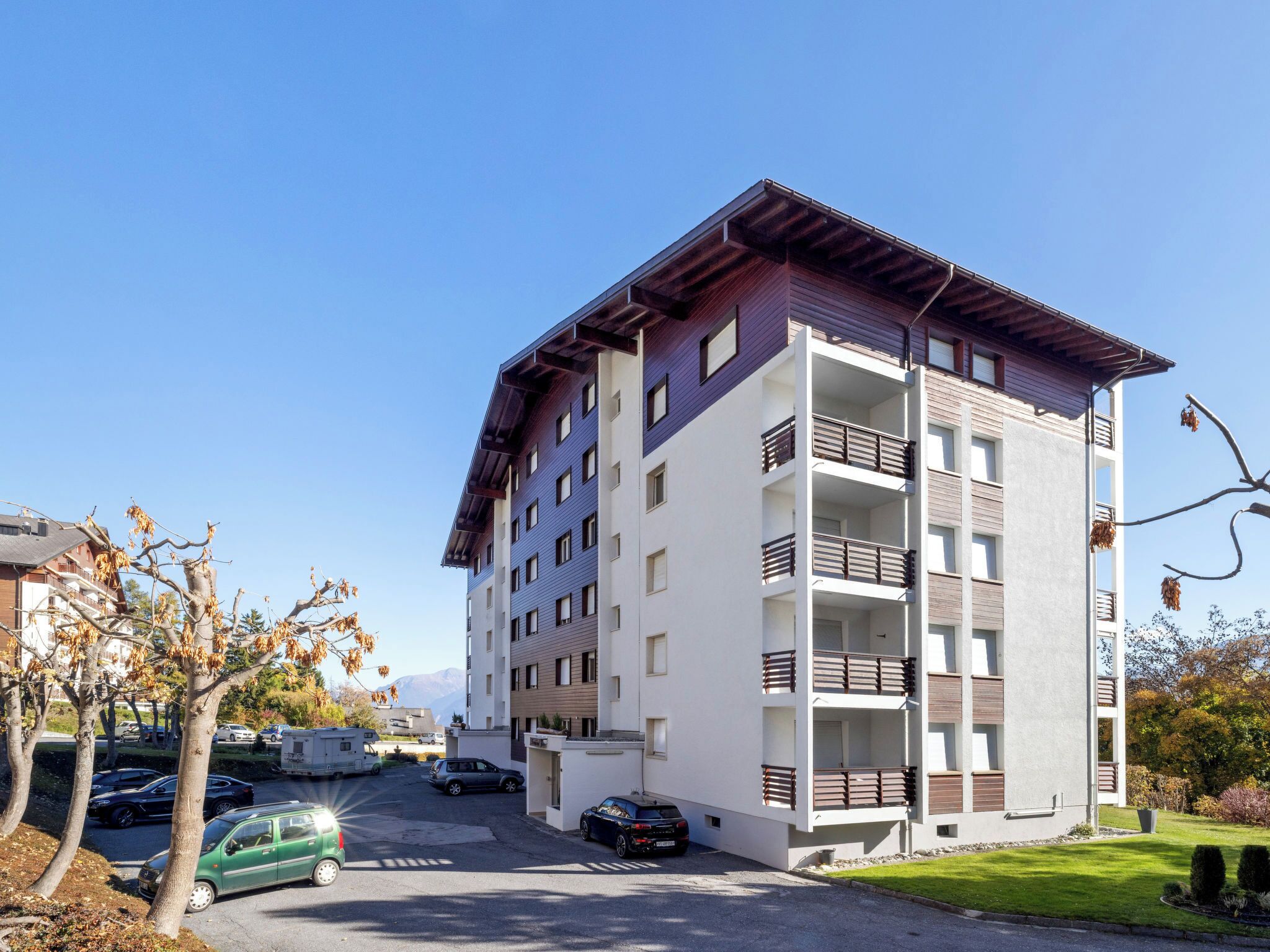 A multi-story residential building with balconies, a parking area, and a clear sky.