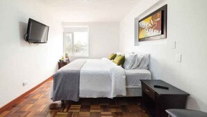 Um quarto com uma cama, uma televisão na parede, uma escrivaninha e uma janela.