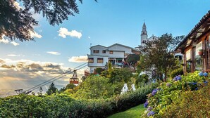 Um hotel numa encosta com um teleférico, jardins exuberantes e a torre de uma igreja ao fundo.