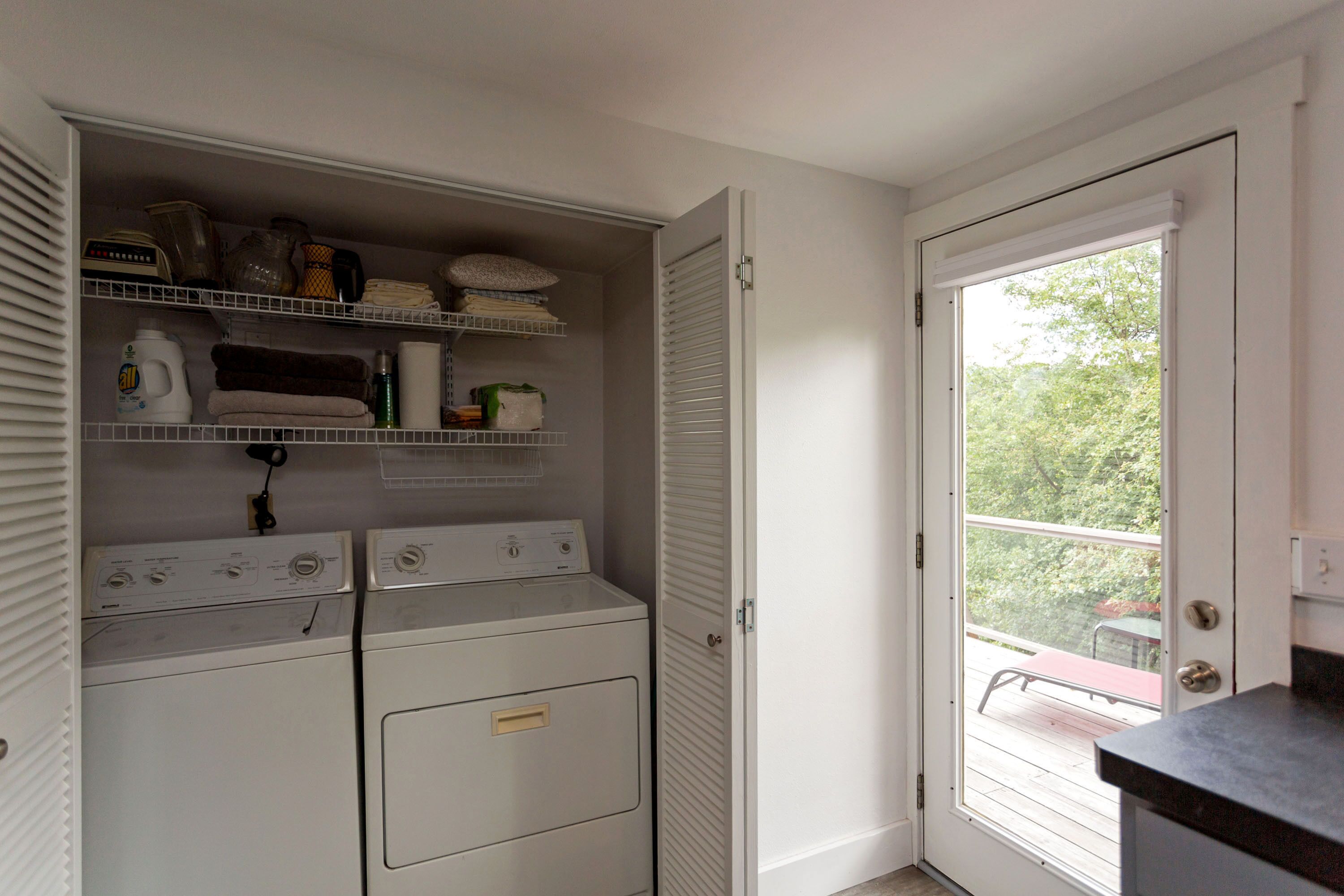 Una sala de lavandería con dos lavadoras, estantes con toallas dobladas y productos de limpieza, y una puerta que da a un balcón exterior.