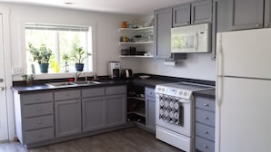 A kitchen with grey cabinets, a white refrigerator, a microwave, a stove, and a window with plants. - Quaint & cozy home in DT Hood River, AMAZING Views off the privacy deck (Hood River)