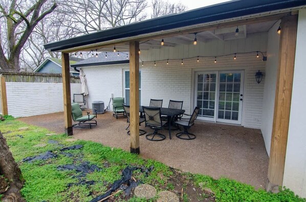 A covered patio with a table and chairs, a covered area with string lights, and a white building in the background.