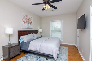 A bedroom with a bed, bedside tables, a ceiling fan, a TV, and a window with blinds.