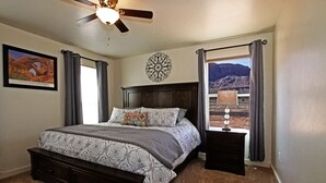 Un dormitorio con cama, mesita de noche, ventilador de techo, ventana con cortinas y decoración en la pared.
