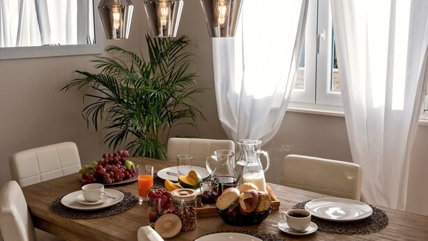 A breakfast table set with fruits, bread, and beverages, surrounded by white chairs and a large plant. - Luxury Villa LeLu with heated saltwater pool suitable for children and pets, with parking, high speed Internet, BBQ (Žrnovnica)