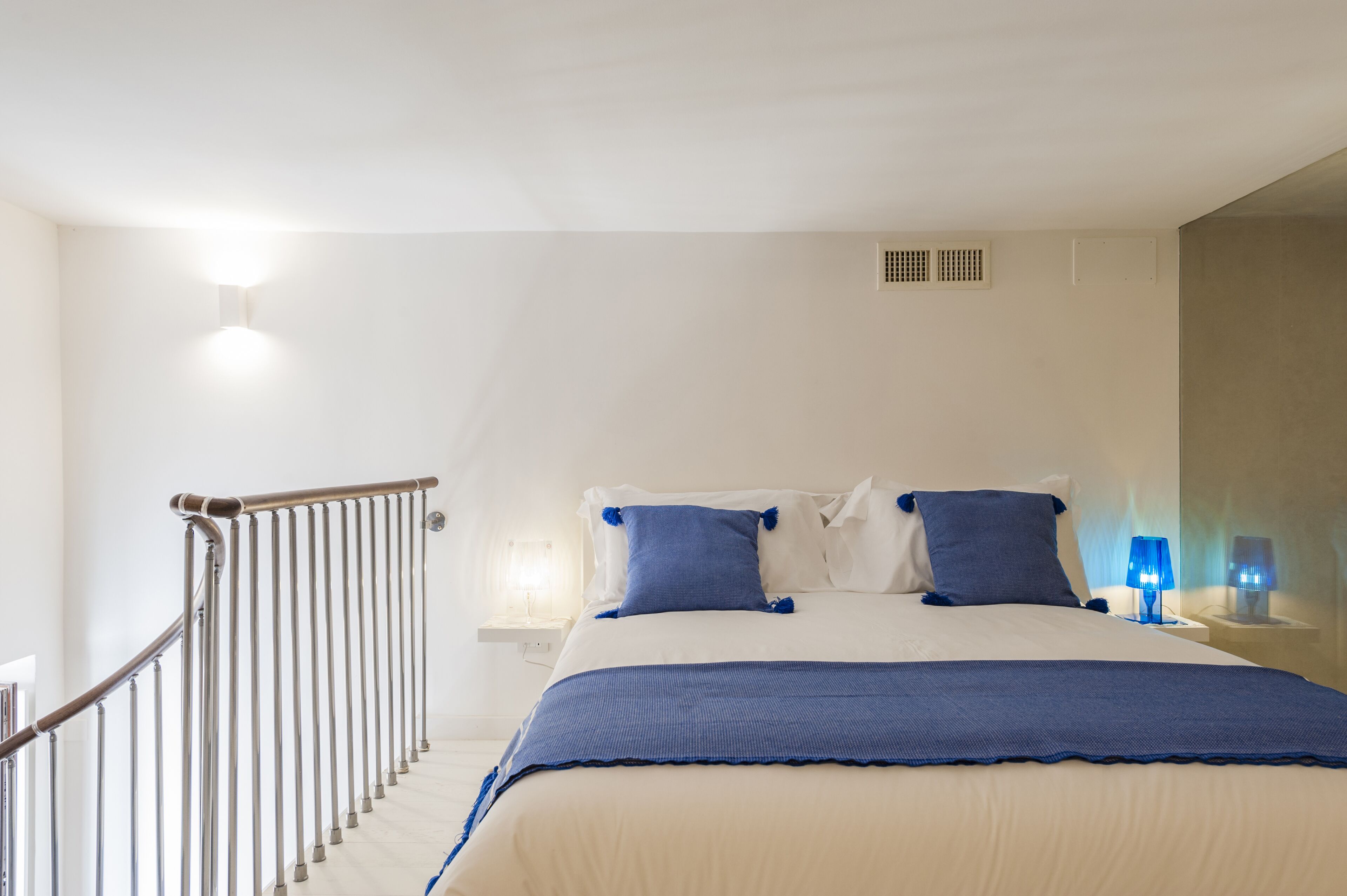 Um quarto com uma cama, uma mesa de cabeceira, uma luminária e o corrimão de uma escada.
