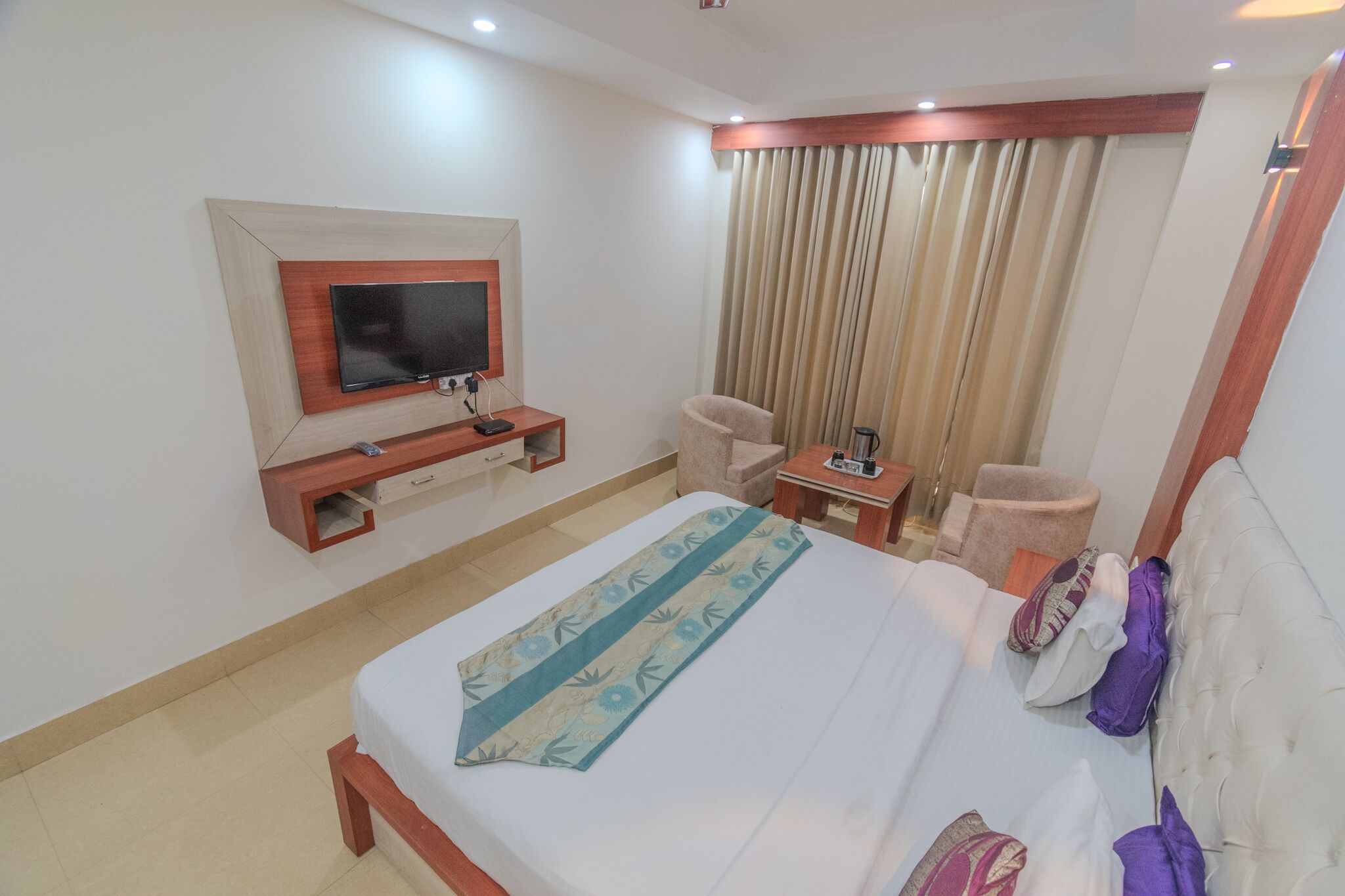 A hotel room with a bed, a TV mounted on the wall, a desk, a chair, and a window with curtains.