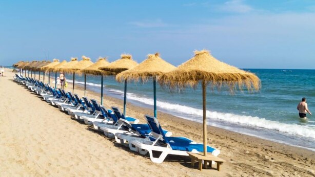 Une plage bordée de rangées de chaises longues bleues et d’ombrelles en chaume.
