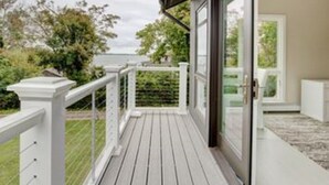 Un espacio al aire libre techado con piso de madera, barandas blancas y puerta de vidrio que da a una habitación.