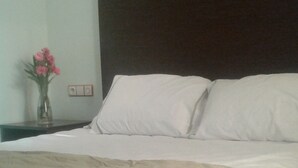 Una cama bien tendida con sábanas blancas y una manta color beige, un cabecero oscuro, y una mesita de noche a la izquierda con un florero de flores rosas.