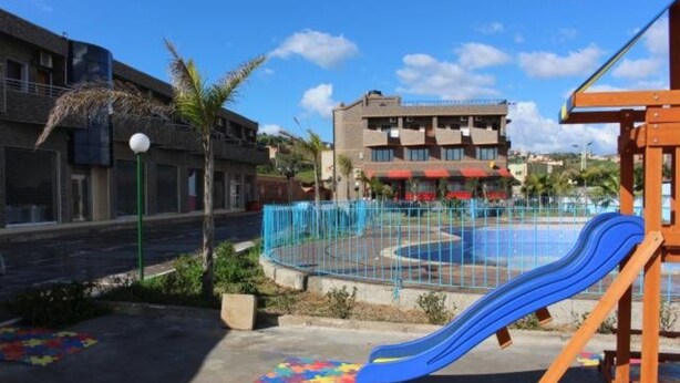 Un parque infantil con un tobogán azul y una zona vallada, rodeado de edificios y un cielo despejado.