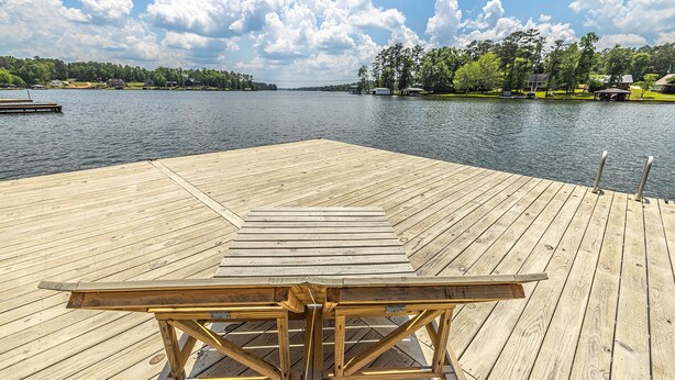 Une jetée en bois avec une table et des chaises, d’où l’on a une vue sur un lac paisible, avec des maisons et des arbres en arrière-plan.