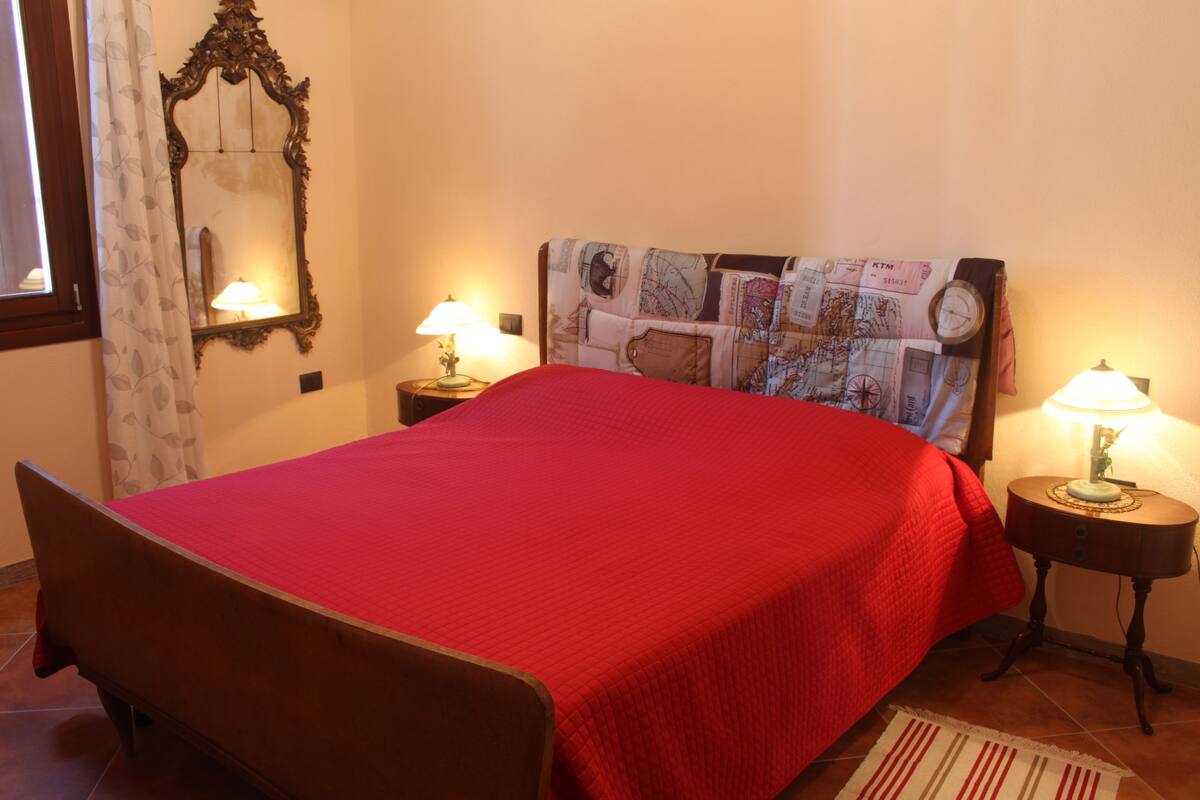 Ein Schlafzimmer mit einem hölzernen Bett, roter Bettwäsche, einem Kopfteil mit Weltkarten-Design, zwei Nachttischlampen, einem Spiegel und einem Fenster mit Vorhängen.