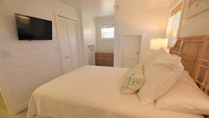 Ein Schlafzimmer mit einem Bett, einem an der Wand montierten Fernseher, einer hölzernen Kommode und einem Fenster mit Jalousien.