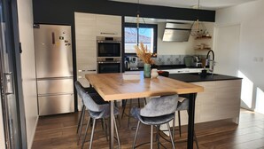 A modern kitchen with a wooden dining table, bar stools, stainless steel appliances, and a window providing natural light. - La Parenthèse Familiale - house with swimming pool between beach and forest (Labenne)