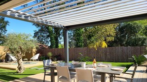 A covered outdoor patio with a table set for dining, surrounded by chairs, and a wooden deck. - La Parenthèse Familiale - house with swimming pool between beach and forest (Labenne)