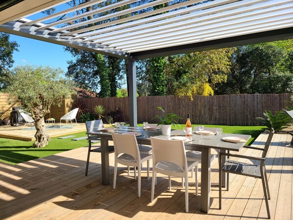 A covered outdoor patio with a table set for dining, surrounded by chairs, and a wooden deck.