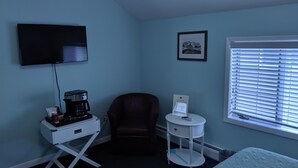 A room with a flat-screen TV mounted on the wall, a coffee maker on a white table, a dark armchair, a small white side table with a remote control, and a window with blinds. - Lake Side Room at The Copper Trout. (Bayfield)