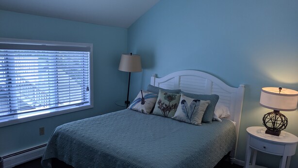 A bedroom with a bed, a bedside table, a lamp, and a window with blinds. - Lake Side Room at The Copper Trout. (Bayfield)
