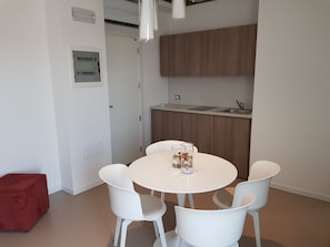 A modern kitchen with white cabinets, a round dining table with four white chairs, and a red ottoman. - Venus Suites (Cervia)