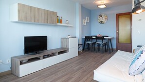 A modern living room with a flat-screen TV, a white sofa, a dining area with a table and chairs, and a wall-mounted shelf with decorative items.