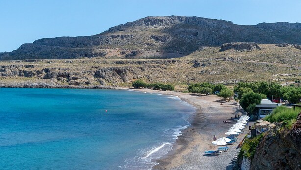 A beach with clear blue water, a rocky mountain in the background, and a row of beach umbrellas and lounge chairs along the shore. - Cretan Minoan Apartments (Sitia)