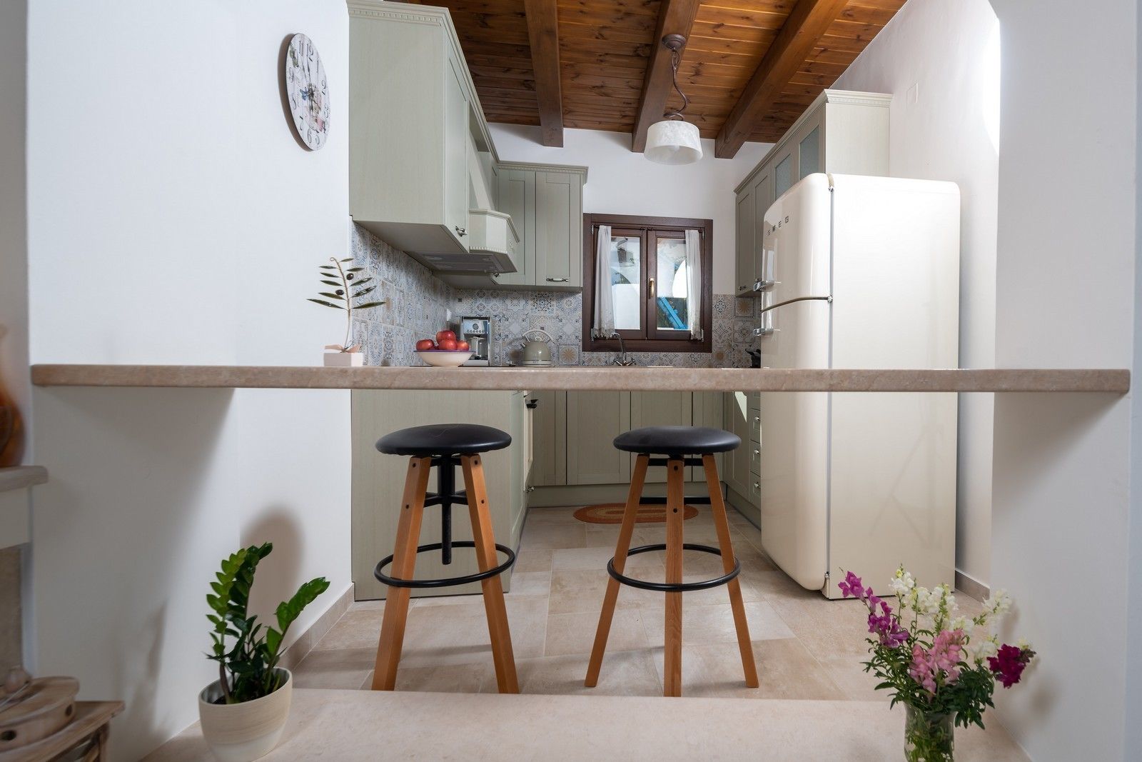 A compact kitchen with a long countertop, two stools, a refrigerator, and a window with curtains.