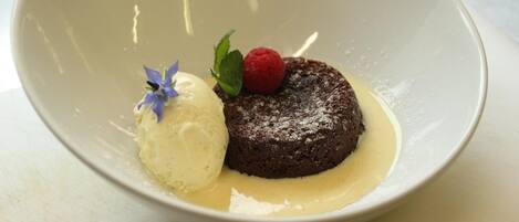 Postre que incluye un pastel de chocolate, adornado con frambuesas y hojas de menta, acompañado de una bola de helado de vainilla en un tazón blanco.