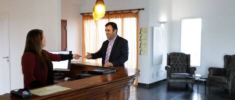 A reception area with a wooden desk, a computer, and a man in a suit. There are two chairs and a small table in the background.