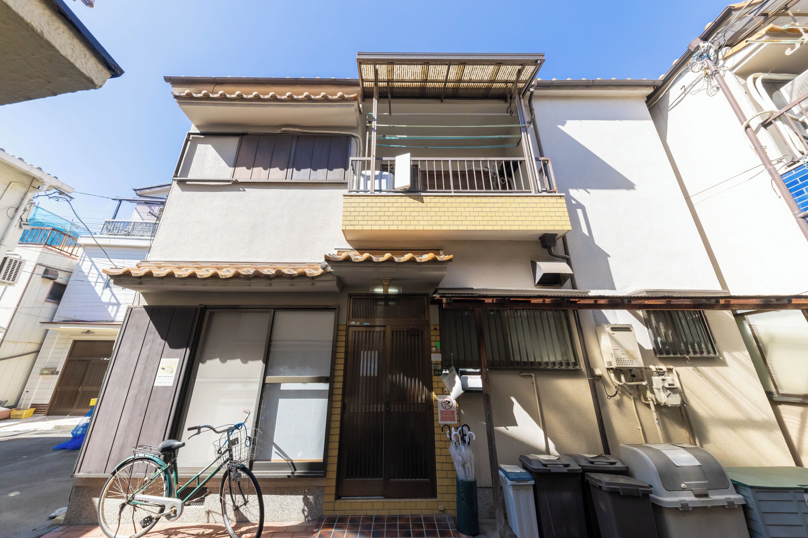 A two-story residential building with a balcony, a bicycle parked outside, and a trash bin.