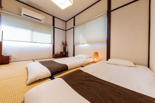 A hotel room with two beds, tatami flooring, a traditional Japanese-style wall, and an air conditioning unit.