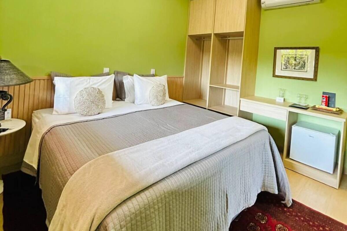Ein Hotelzimmer mit Bett, Nachttisch mit Lampe, Kleiderschrank, Schreibtisch mit Stuhl und kleinem Kühlschrank.