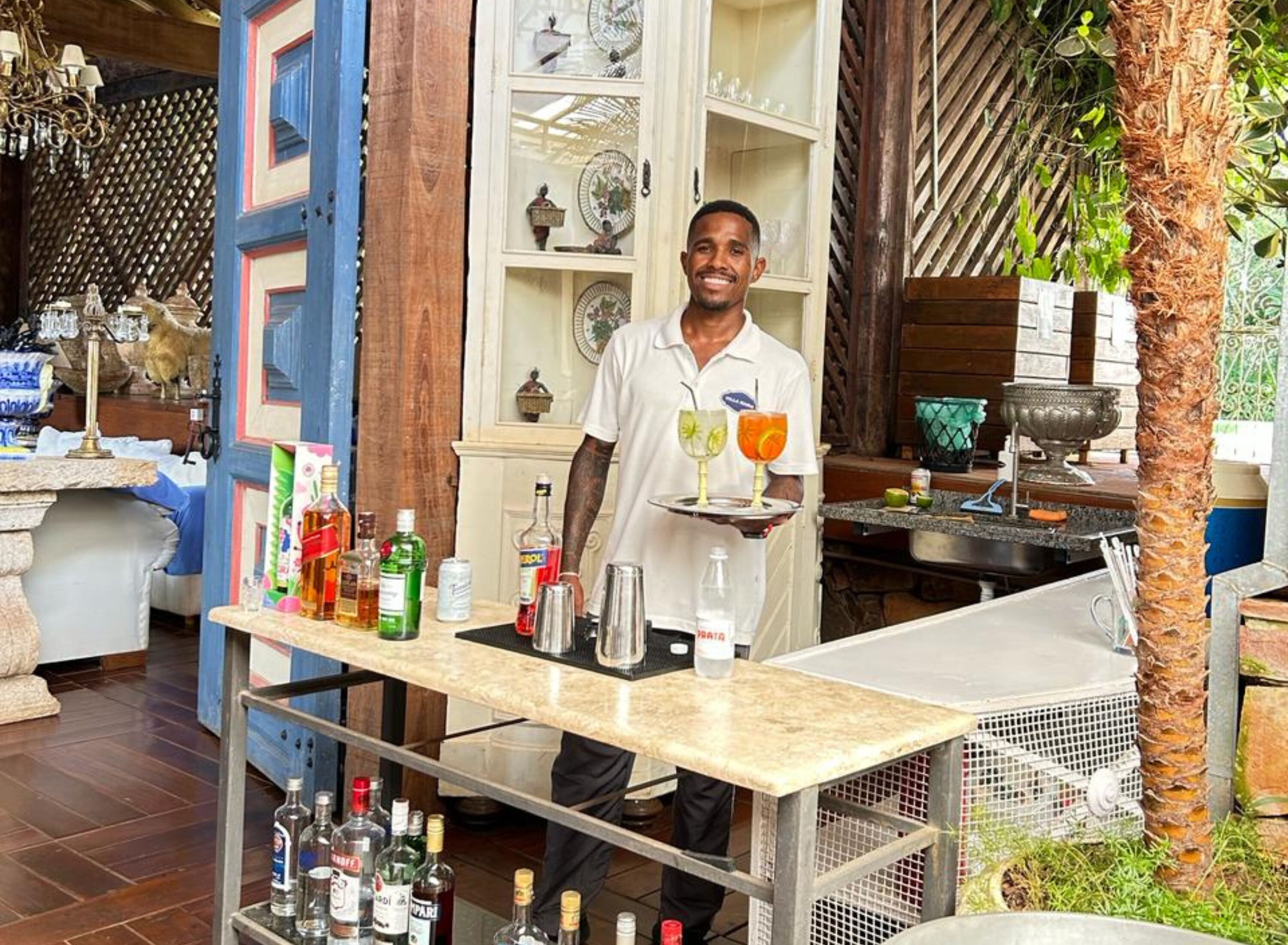 A bar area with a bartender holding a tray of drinks, shelves with bottles, and a decorative wall.