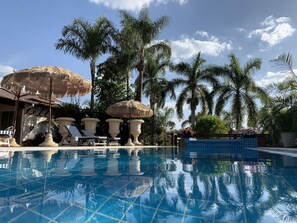 A swimming pool with clear blue water, surrounded by palm trees and thatched umbrellas.