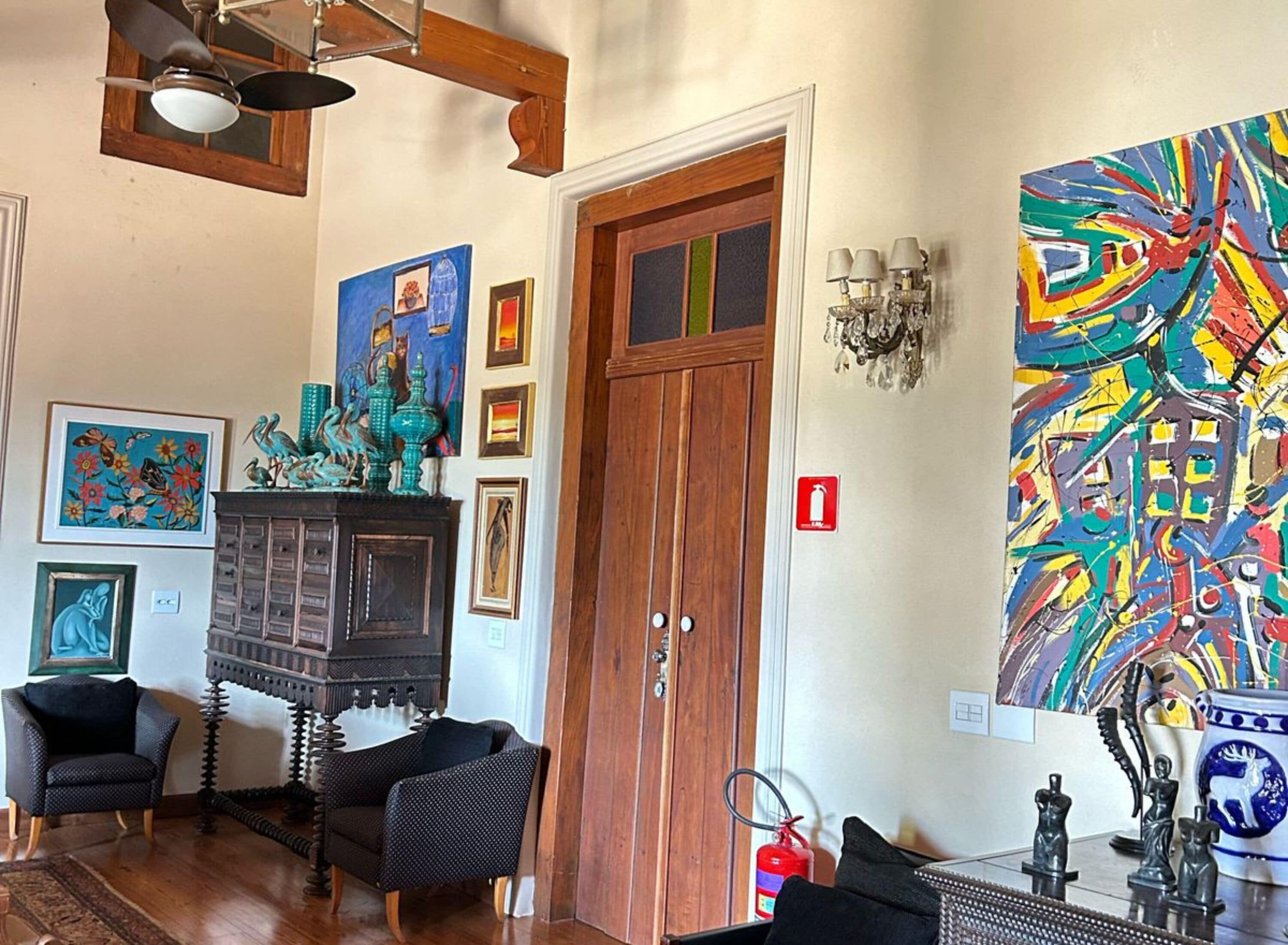 A room with a wooden door, a fireplace, two black chairs, a colorful abstract painting, and a chandelier.