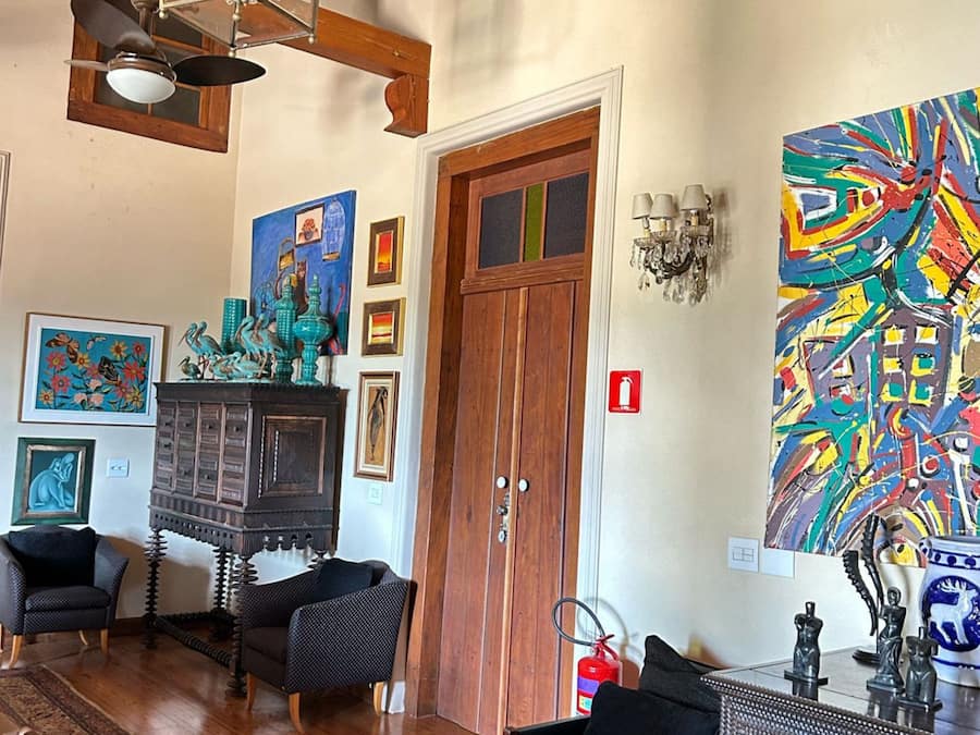 A room with a wooden door, a fireplace, two black chairs, a colorful abstract painting, and a chandelier.
