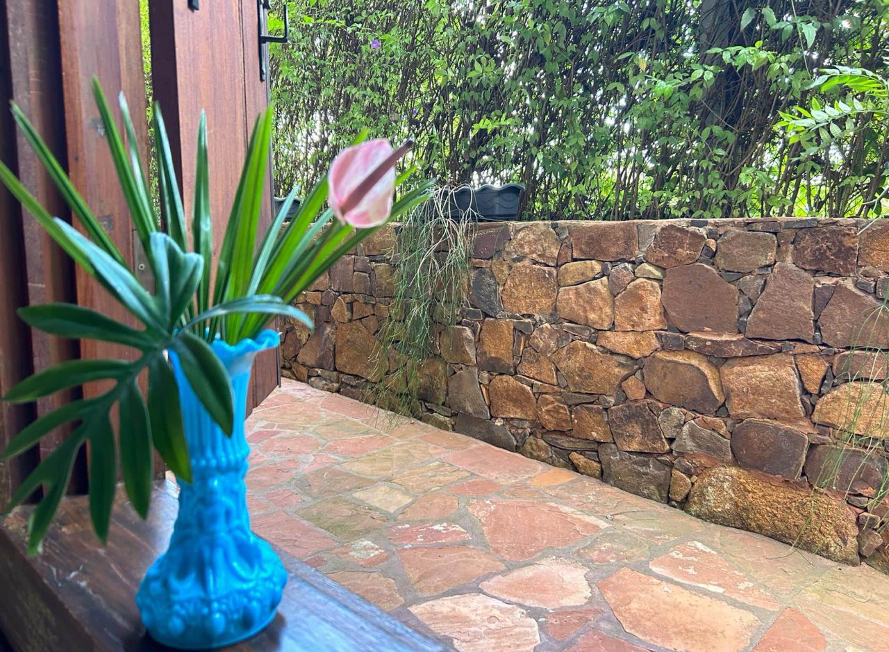 A blue vase with a plant on a stone-tiled ledge, a stone wall, and lush greenery in the background.