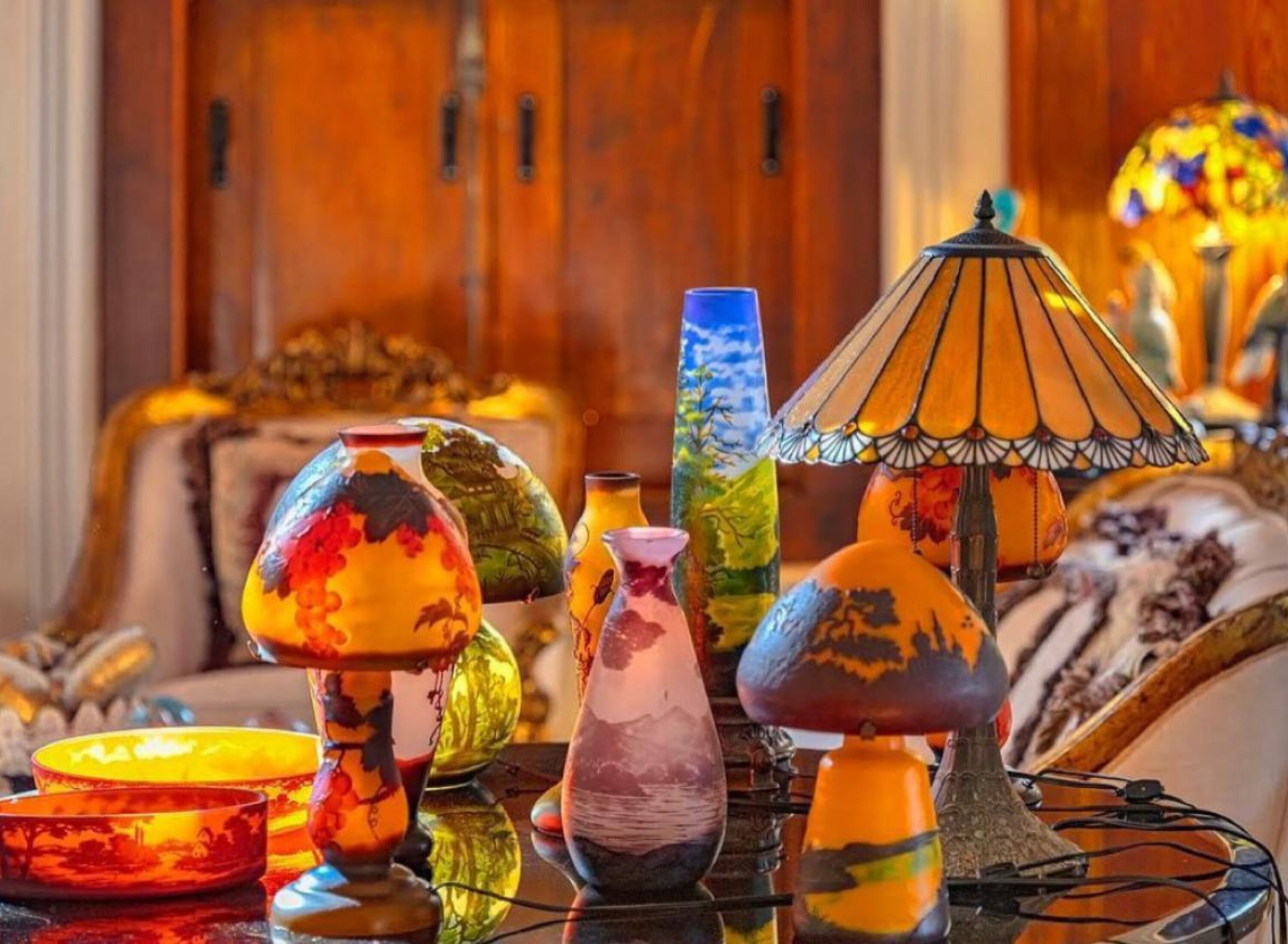 Diversos copos e taças coloridos, além de um abajur sobre uma mesa em um ambiente acolhedor com móveis de madeira.