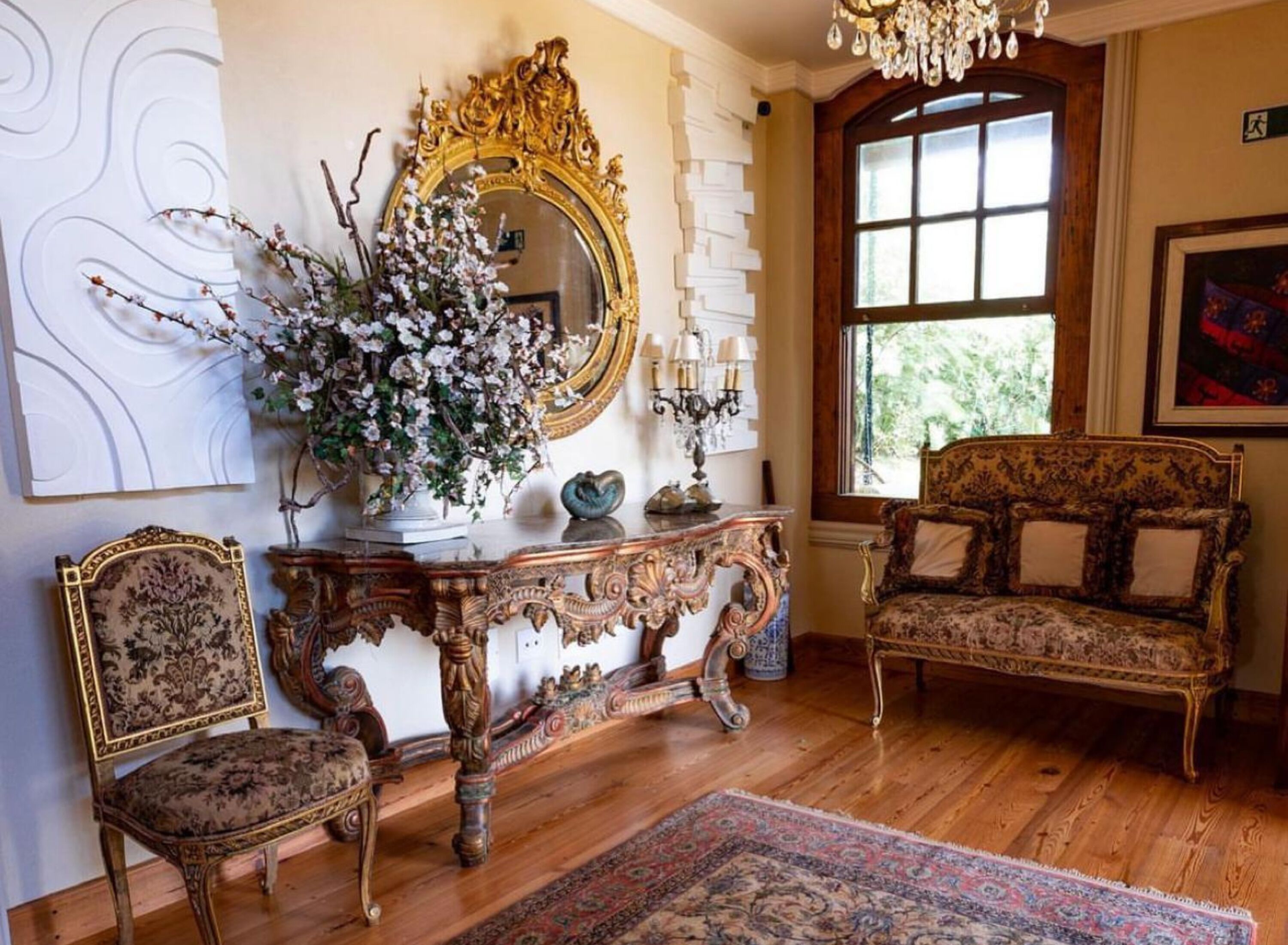 A room with a large ornate mirror, a floral arrangement, a vintage sofa, and a patterned rug.
