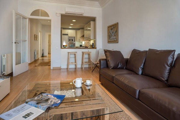 A living room with a brown leather sofa, a glass coffee table with a magazine, and a kitchen in the background.