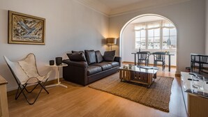 Una sala de estar moderna con un sofá de cuero marrón, un sillón blanco, una mesa de centro de vidrio y un comedor con mesa y sillas.