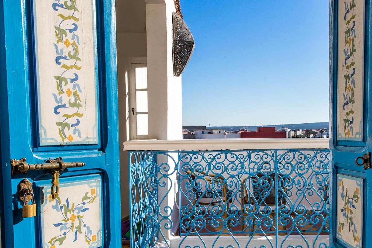 An open blue door with decorative tiles, leading to a balcony with a blue wrought iron railing and a view of the cityscape.