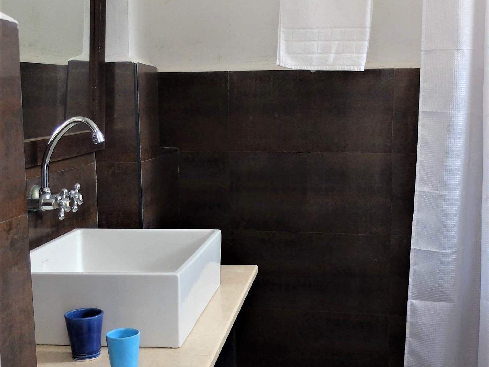 Ein Badezimmer mit einem weißen Waschbecken, dunkelbraunen Fliesen, einem Spiegel und einer blauen Tasse auf der Ablagefläche.
