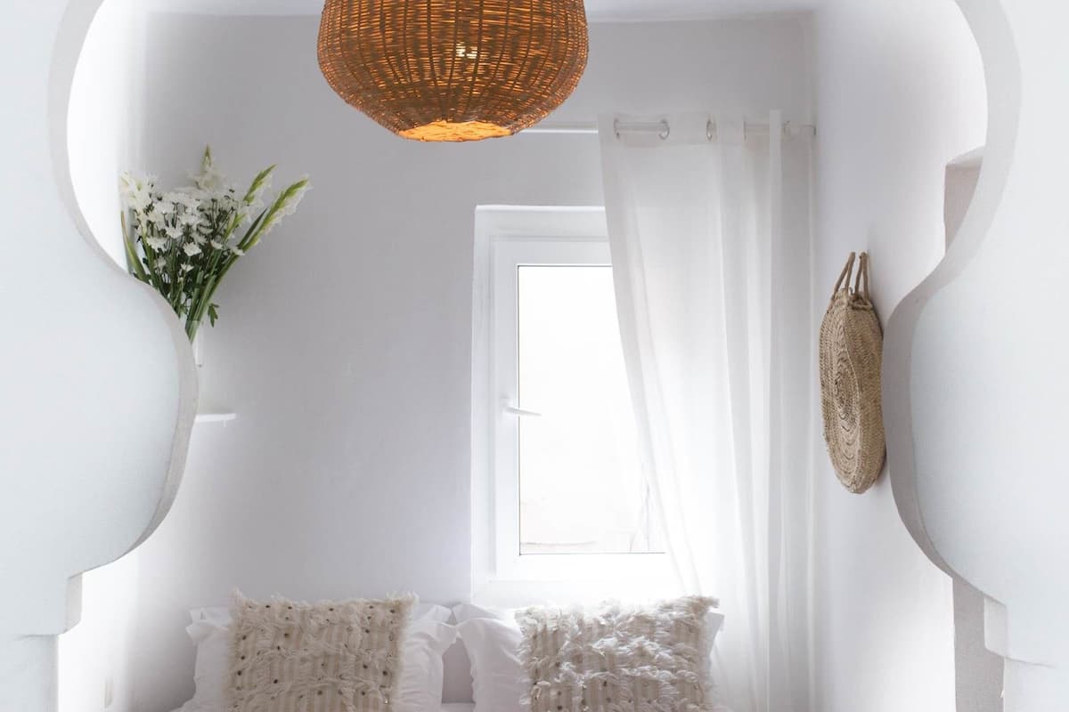 A white room with a bed, a wicker light fixture, a window with sheer curtains, and a wall-mounted decorative item.