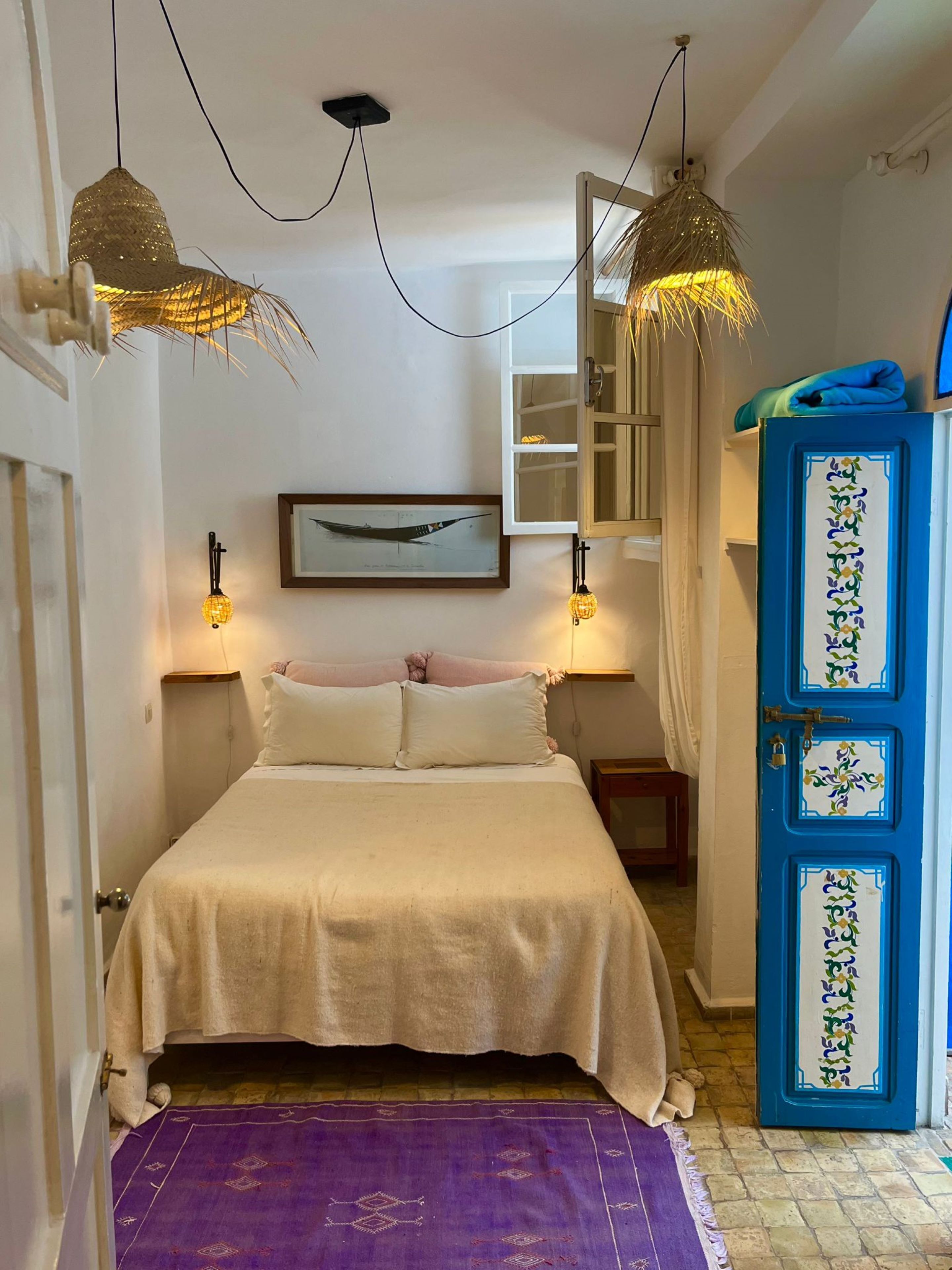 A bedroom with a bed, two hanging lamps, a framed picture on the wall, a blue cabinet, and a purple rug.