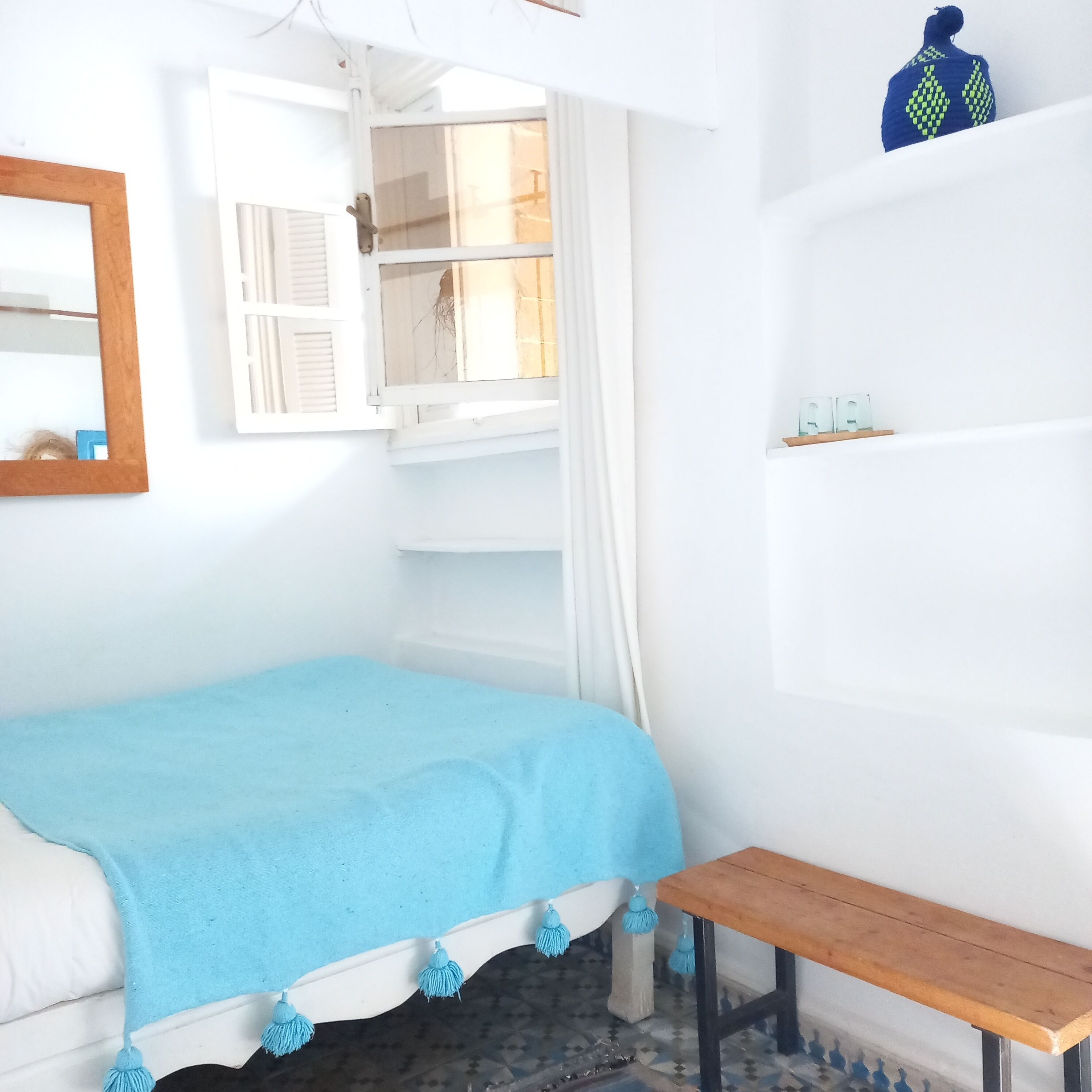 Quarto com cama, janela, um banco de madeira e uma estante com objetos.