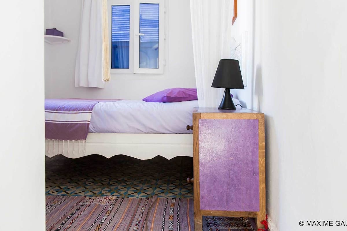 A bedroom with a bed, a nightstand with a lamp, a window with curtains, and a patterned rug.