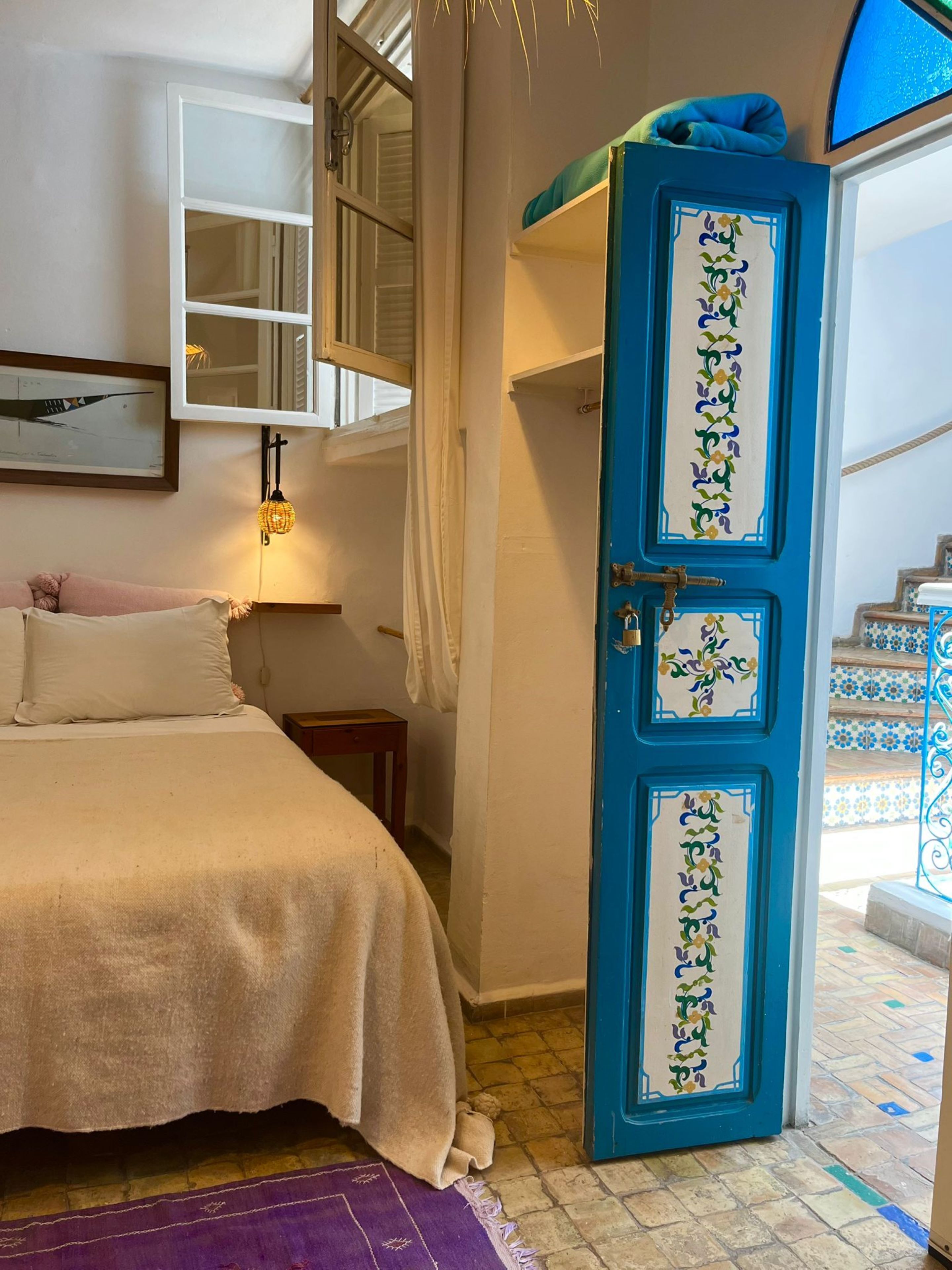 A bedroom with a bed, a nightstand, a window with curtains, and a blue door with decorative panels.