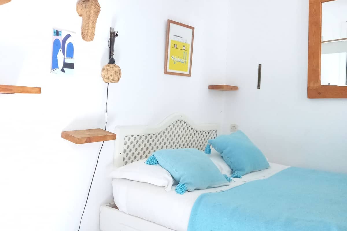 A bedroom with a bed, a hanging lamp, a framed picture, and a small shelf.