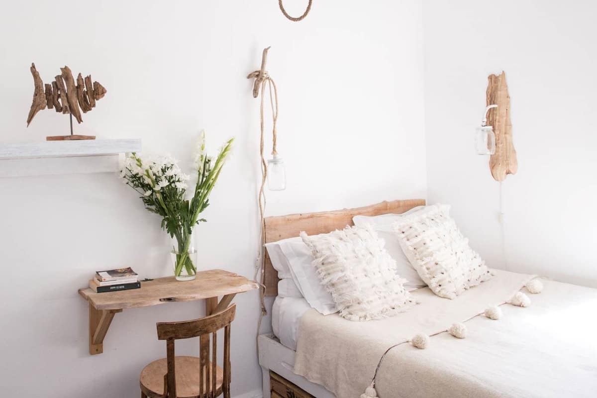 A bedroom with a wooden bed, white pillows, a bedside table with a vase of flowers, and a rustic wall decor piece.